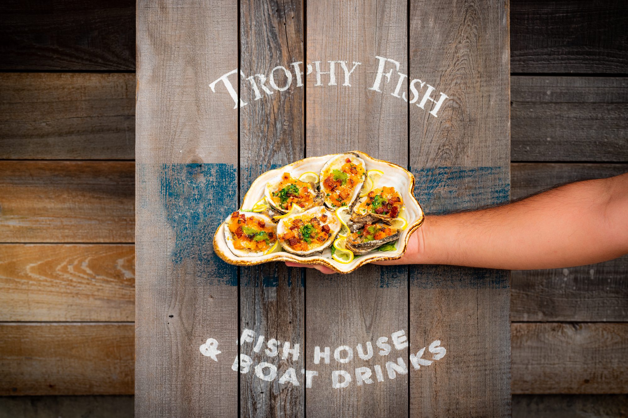 A hand holding an order of fish tacos in front of the weathered-wood Trophy Fish wall lettered “Fish House & Boat Drinks.”