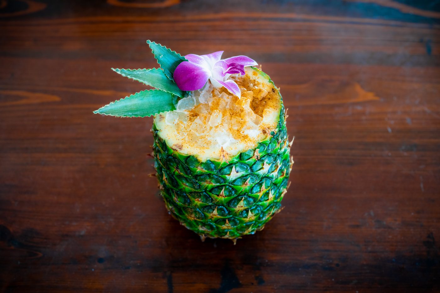 A frozen cocktail served in a carved pineapple with a palm-leaf and orchid garnish.